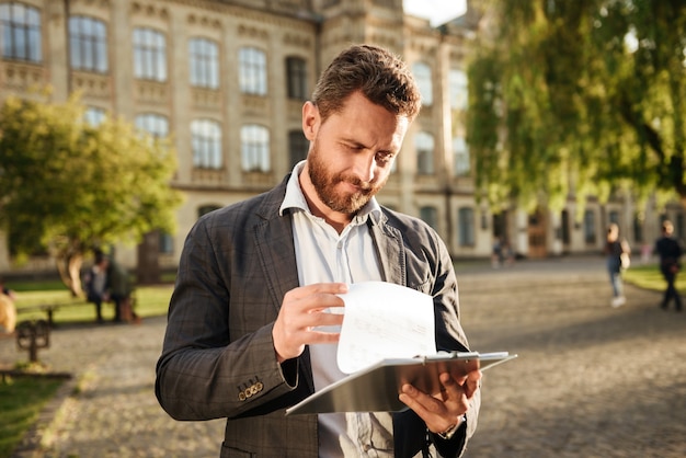 serious man holding clipboard and examining documents, while standing in front of old building in park