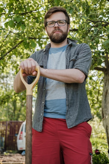 Serious man is leaning on the handle of a shovel and looking at the camera in the garden