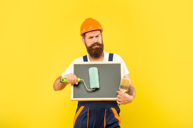 Serious man renovator in helmet and work clothes hold paint roller brush and blackboard