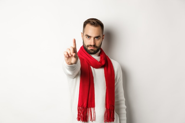 Serious man saying no, showing stop sign, shaking finger in decline, rejection sign, prohibit action, standing in winter sweater and red scarf against white background