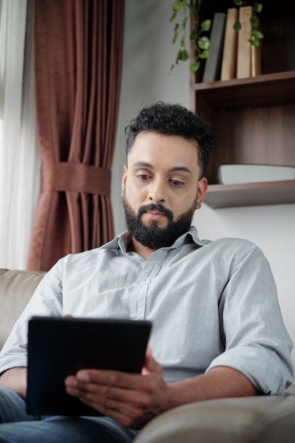 Serious man sitting in armchair and reading book or document on tablet computer