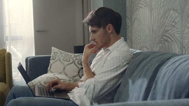 Serious man sitting on couch with computer Focused guy working on laptop inside