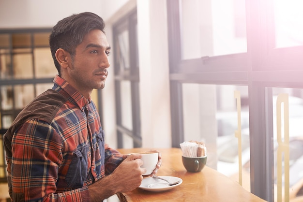 深刻な考え方をしている男性と朝の朝食にコーヒーを飲む窓または屋内カフェでのビジョンレストランでコーヒーやお茶を飲むカップや飲み物を飲むと思った若い男性