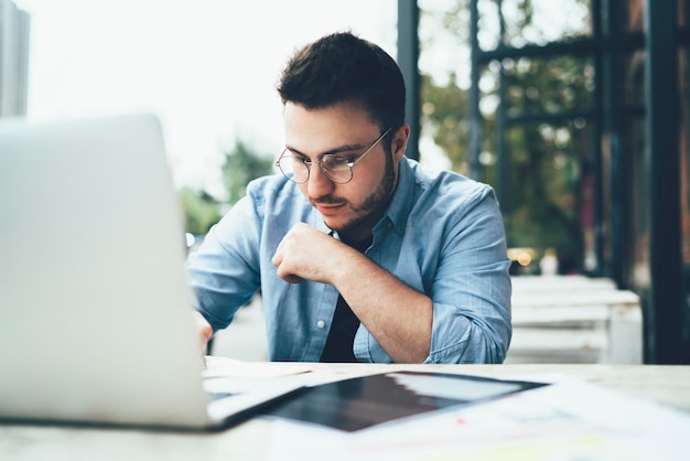 Serious man thoughtfully looking at documents working in cafe with laptop