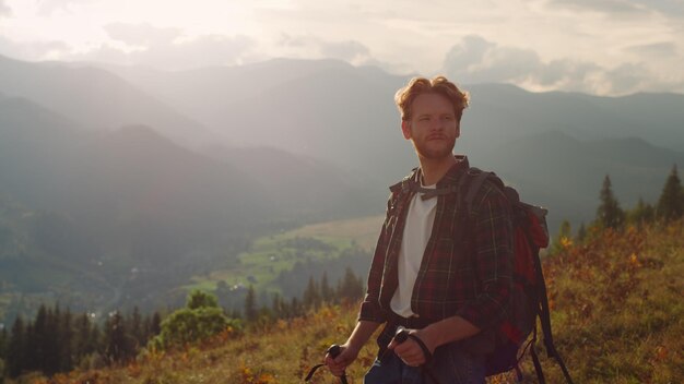 Photo serious man travel mountains landscape hipster tourist using trekking poles closeup relaxed backpacker discover walk green hill at sunset redhead guy hiking on expedition active leisure concept