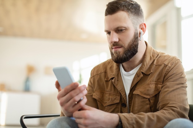 Serious man using smartphone wearing earbuds earphones at home