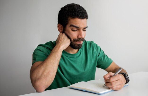 Foto uomo serio con la barba con una maglietta verde che scrive su un taccuino appoggiato al mento