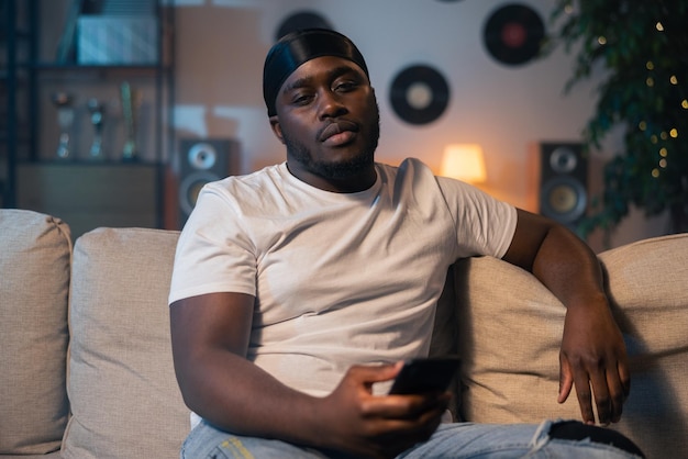 A serious man with dark skin is resting on the couch in the evening looking at the camera