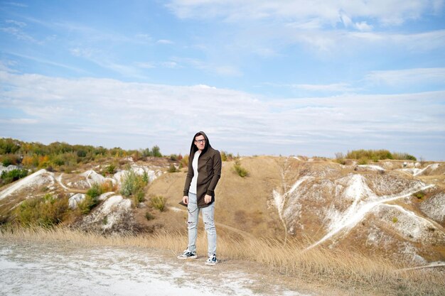 Serious man with glasses in jeans tshirt and cape stands in hilly terrain Adult male with eyeglasses in hood looks away thoughtfully in countryside