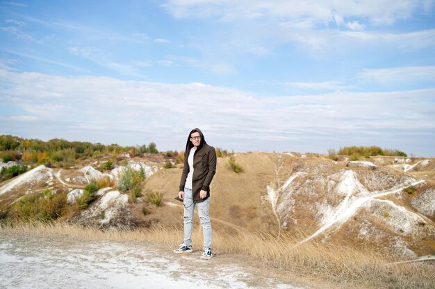 Uomo serio con occhiali in jeans, maglietta e mantello si trova in un terreno collinare maschio adulto con occhiali in cappa distoglie lo sguardo pensieroso in campagna