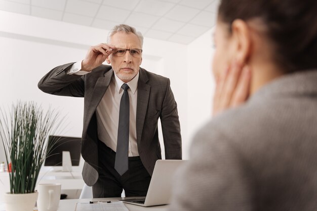 Photo serious man in years wearing costume pressing lips while touching his glasses