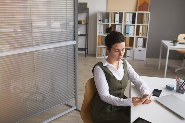 Serious manager sitting at her workplace with mobile phone and working online