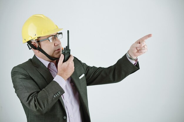 Serious mature building contractor or engineer in work helmet and glasses using walkie-talkie while explaining project plan to worker