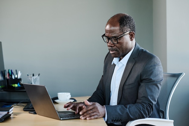 Serious mature businessman in formalwear and eyeglasses using laptop