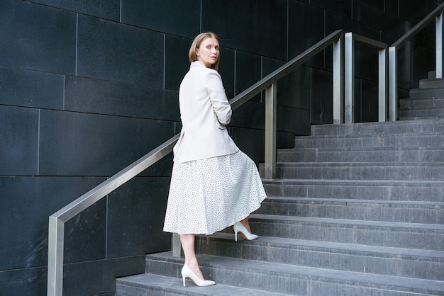 Serious mature businesswoman in elegant dress standing on stairs in front of an office building coac...