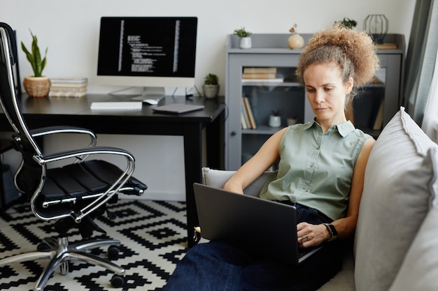 Serious mature businesswoman sitting on sofa and working on laptop in the room