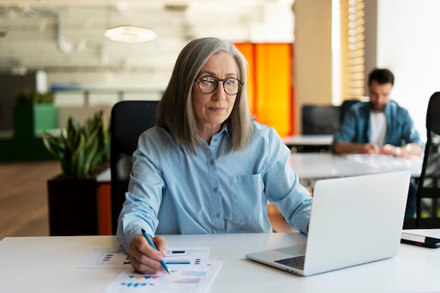 Серьезная зрелая деловая женщина, использующая портативный компьютер, делает заметки, рабочий проект в современном офисе. 60-летняя женщина-финансист, работающая с документами на рабочем месте. Успешная деловая карьера.