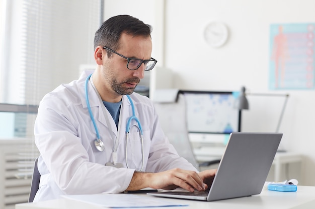 Serious mature doctor in eyeglasses sitting at the table and typing on laptop computer at office