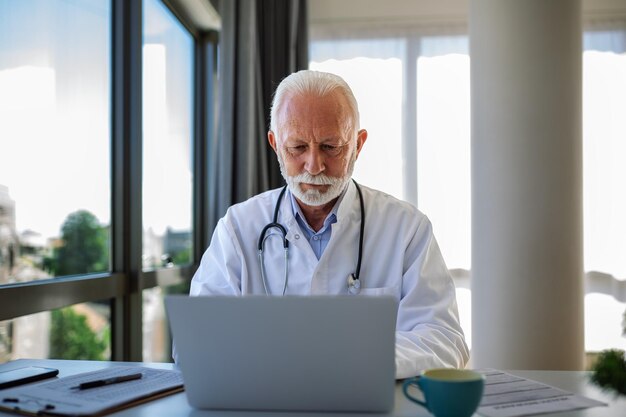 Serious mature doctor using laptop and sitting at desk Senior professional medic physician wearing white coat and stethoscope working on computer at workplace