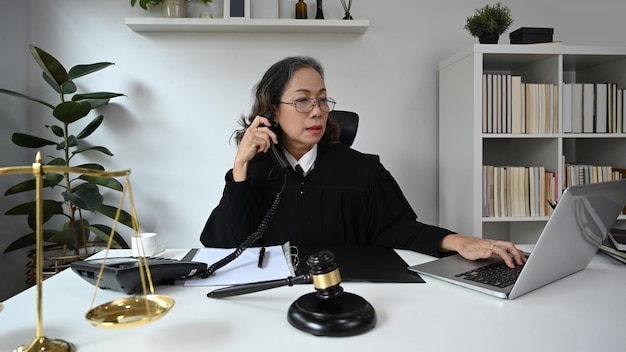 写真 彼女のオフィスでラップトップを使用してガベルハンマー槌でテーブルに座っている深刻な成熟した女性裁判官または弁護士またはローブガウンの制服