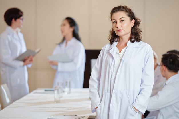 Serious mature female pharmacologist in labcoat standing at table in boardroom and looking at camera