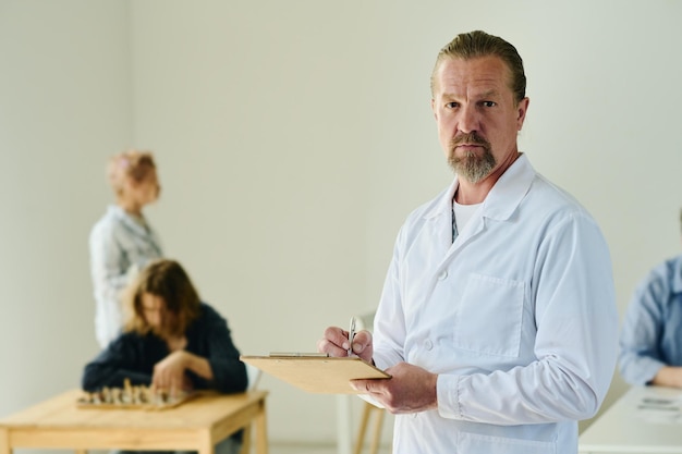 Serious mature male psychiatrist in lab coat looking at camera in mental hospital