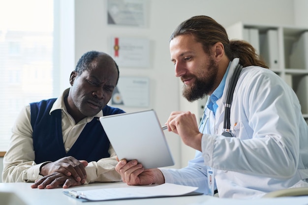 Фото Серьезный взрослый пациент смотрит на экран планшета и слушает врача