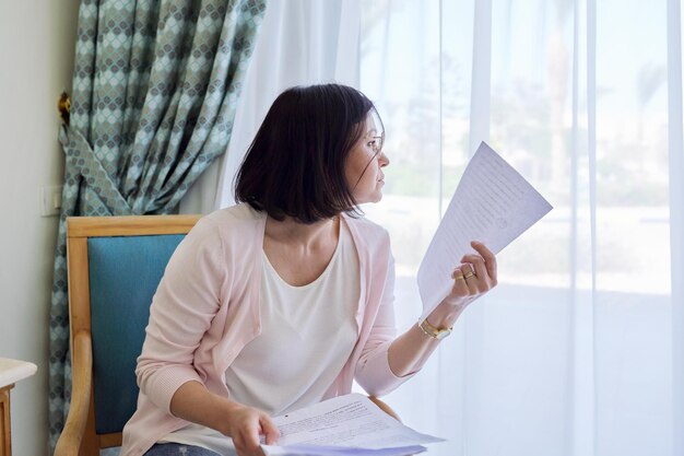 Serious mature woman reading papers documents sitting in
armchair at home