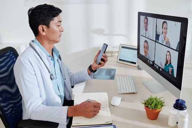 Serious middle-aged Asian doctor having online conference with colleagues and taking notes in planner