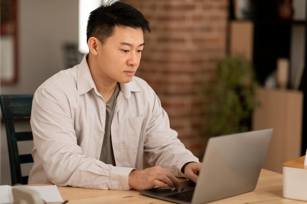 Серьезный азиат средних лет, использующий портативный компьютер, печатающий на клавиатуре во время работы в Интернете, сидя в домашнем офисе