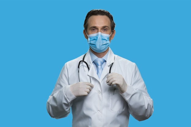 Serious middle-aged doctor is wearing stethoscope. Physician in a white coat and protective mask isolated on blue background.