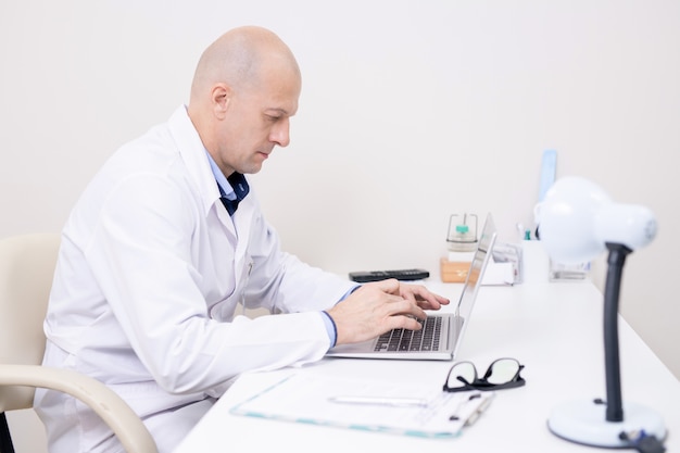 Serious middle aged doctor in whitecoat concentrating on laptop work while sitting by desk and typing in clinics