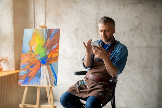 Photo serious middle aged painter in apron and casualwear scrolling in smartphone while sitting on chair in studio