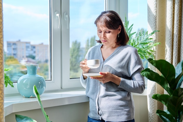 Серьезная женщина средних лет с чашечкой кофе смотрит в окно.