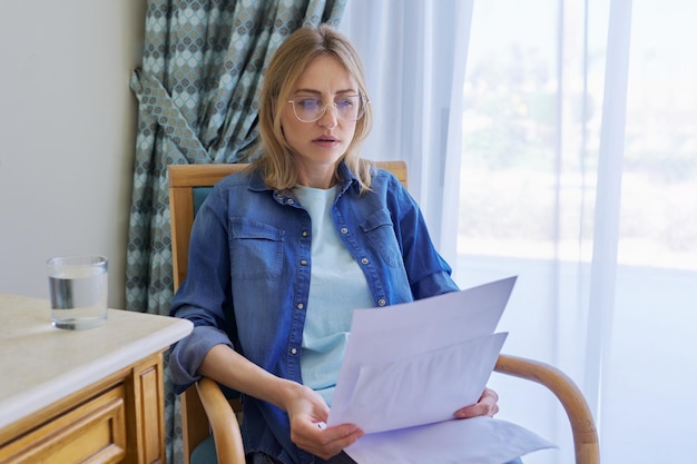 Serious middle aged woman with glasses reading papers sitting at home in chair