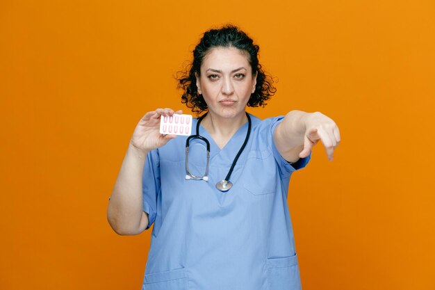 Serious middleaged female doctor wearing uniform and stethoscope around her neck showing pack of capsules looking and pointing at camera isolated on orange background