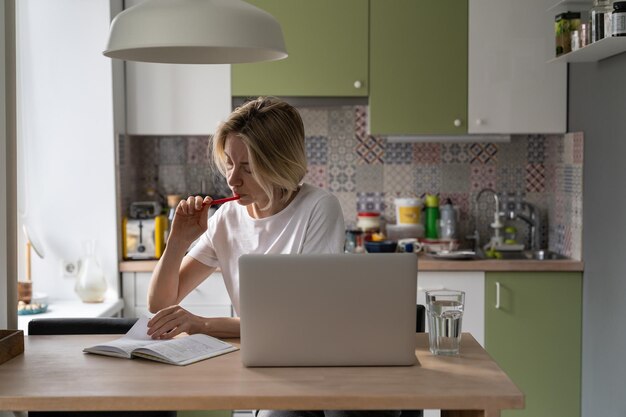 Serious middleaged woman sits in kitchen making notes about job vacancies in paper pad