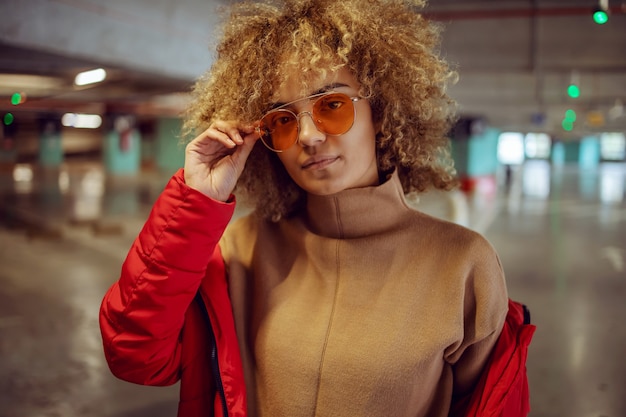 Photo serious mixed race hip hop girl in jacket standing in garage and adjusting sunglasses while looking at camera.