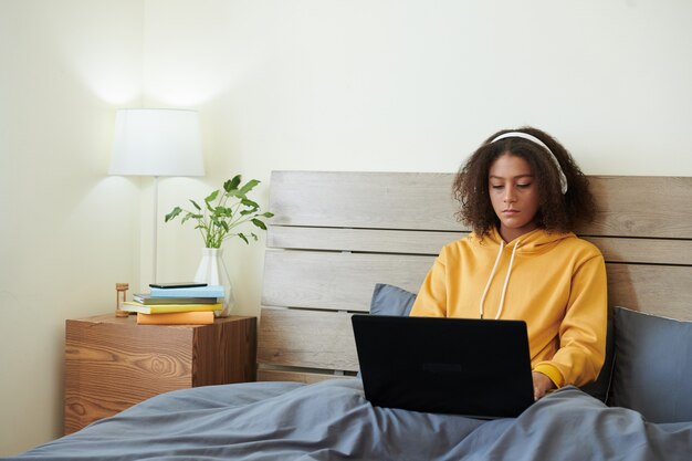 Serious mixed race teenage girl in headphones sitting under duvet in bed and using laptop at home