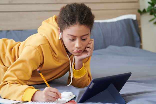Серьезная девочка-подросток смешанной расы в желтой толстовке с капюшоном, лежа на кровати и используя планшет, делая заметки в рабочей тетради