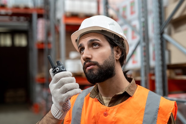 Foto operaio serio di magazzino di razza mista in elmetto protettivo bianco che trasmette un messaggio al collega tramite walkie-talkie