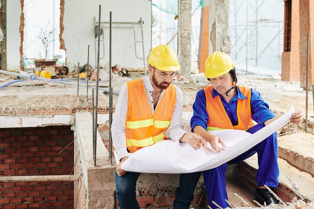 Serious multi-ethnic builders sitting in room of builing under construction and discussing blueprint