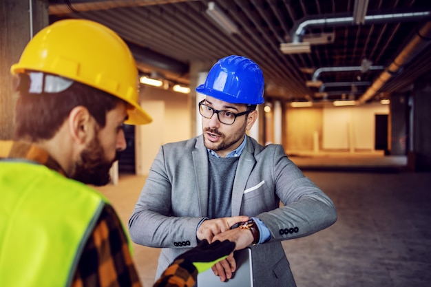 Serious nervous supervisor arguing with his worker and pointing at wristwatch. Work must be done on time with no excuses. Building in construction process interior.