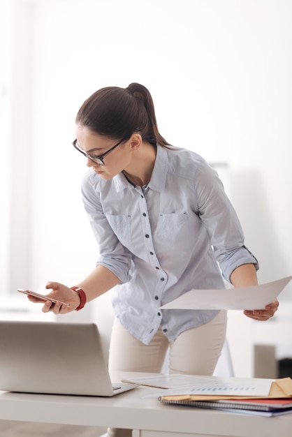 Serious office worker holding phone in right hand keeping document in left one while looking at her device