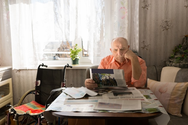Серьезный старик, серьезно читающий новости таблоидов, отдыхая в гостиной со своим креслом-каталкой сбоку.