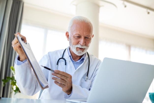 Serious old mature professional male doctor using laptop computer in hospital office having medical webinar training writing in healthcare report consulting patient online at telemedicine meeting