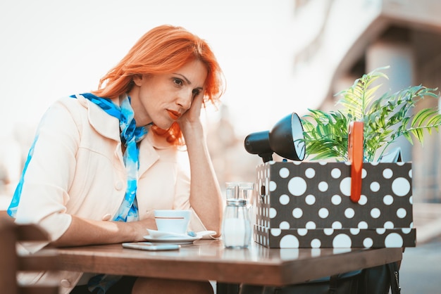 Serious pensive businesswoman sitting in a street cafe with a box full of her personal belongings from the office just after she got fired.