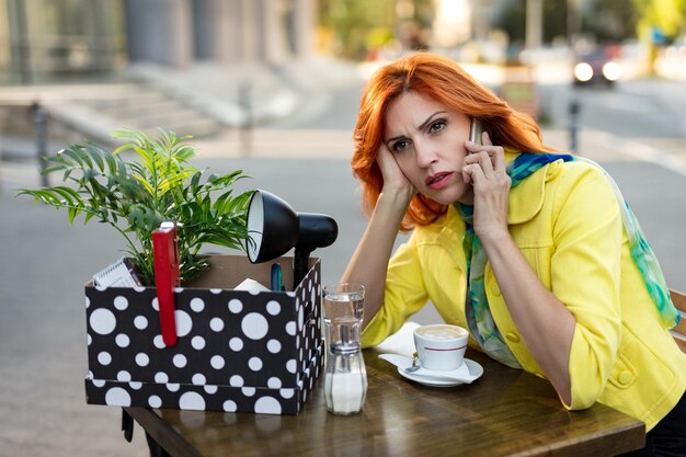 Серьезная задумчивая деловая женщина сидит в уличном кафе с коробкой, полной вещей из офиса, когда он уволился, и разговаривает по смартфону.