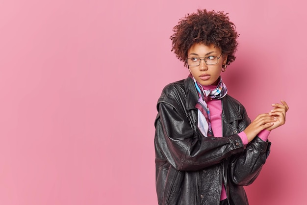 Serious pensive curly haired young woman wears leahter jacket kerchief around neck keeps hands together focused away isolated over pink background with copy space for your promotional content