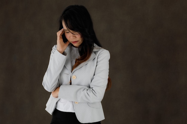 Serious pensive young Asian businesswoman in classy outfit touching forehead and close eyes thoughtfully against dark gray background with empty space